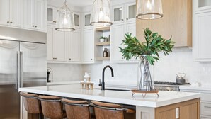 Una cocina moderna con armarios blancos, un refrigerador de acero inoxidable, una isla central con taburetes de bar, luces colgantes y un jarrón con hojas verdes.