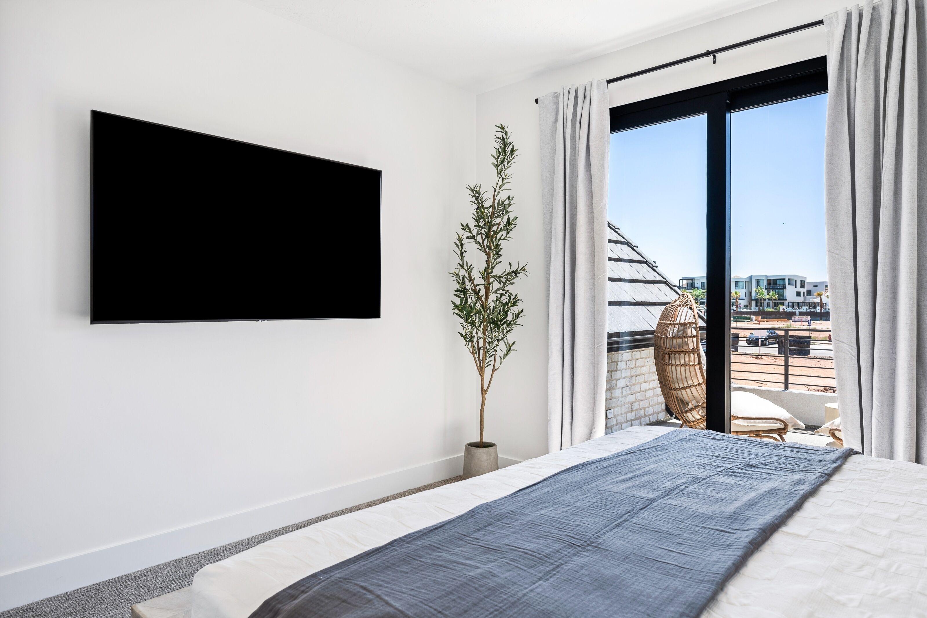 Camera moderna con un letto grande, una TV a parete, una pianta in vaso e vista sulla città attraverso un'ampia finestra.