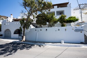 A white building with a tree in front, a white wall with a sign, and a clear sky. - Zisis Pension (Mykonos)