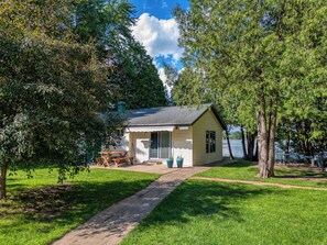 A small, single-story cabin with a porch, situated in a grassy area with trees, near a body of water.