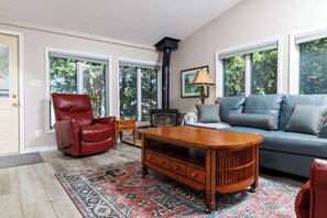A living room with a sofa, a red leather chair, a wooden coffee table, a fireplace, and large windows with a view of trees.