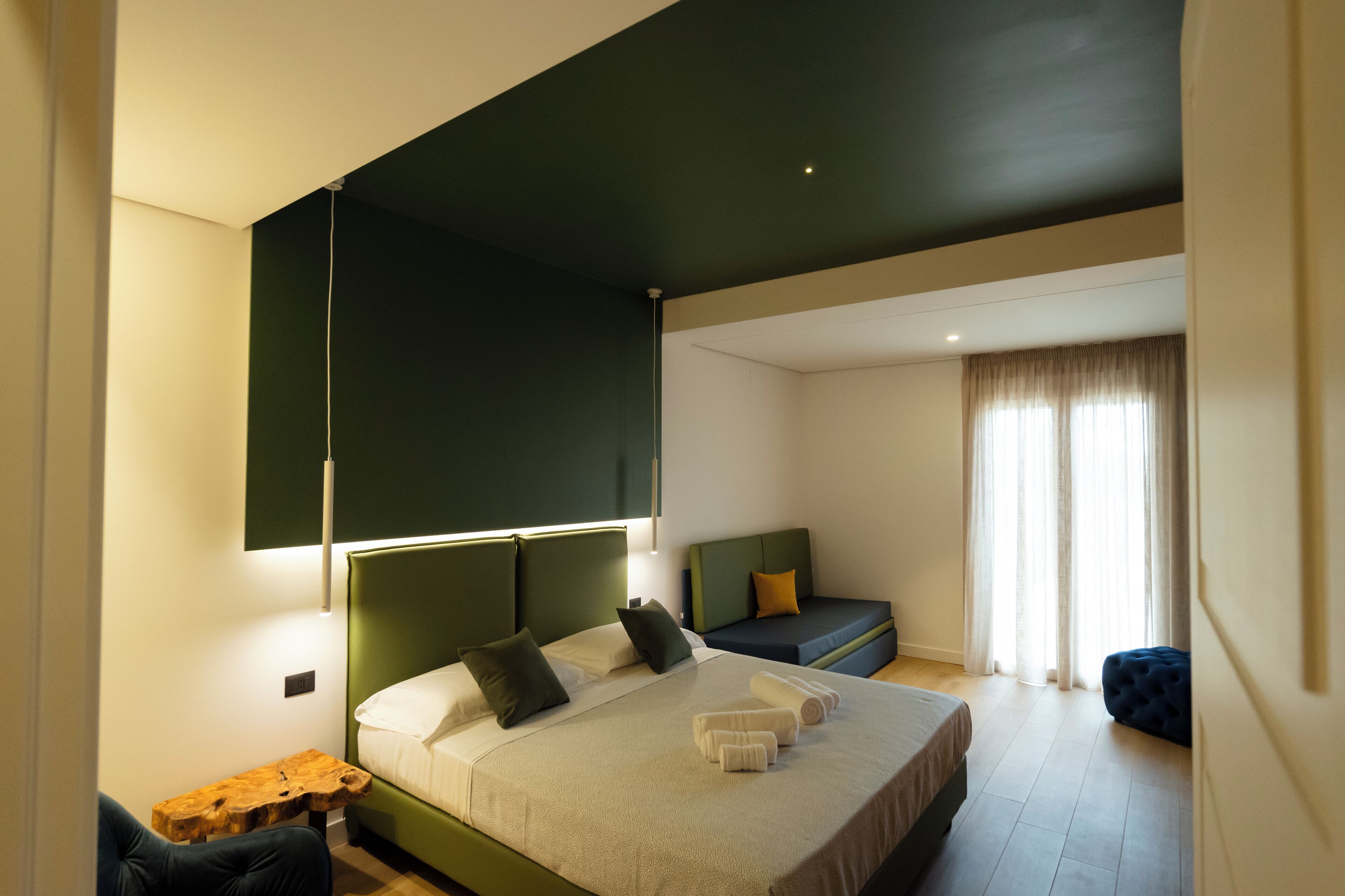 Quarto de hotel moderno com uma cama grande, um sofá pequeno, uma mesa de cabeceira de madeira e uma janela com cortinas.