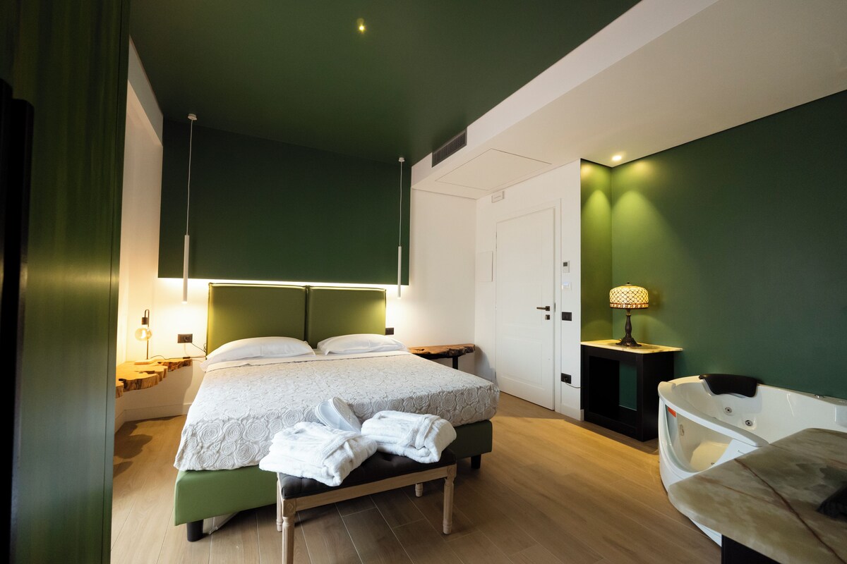 A modern hotel room with a bed, bedside tables, a bench, and a small table with a lamp.