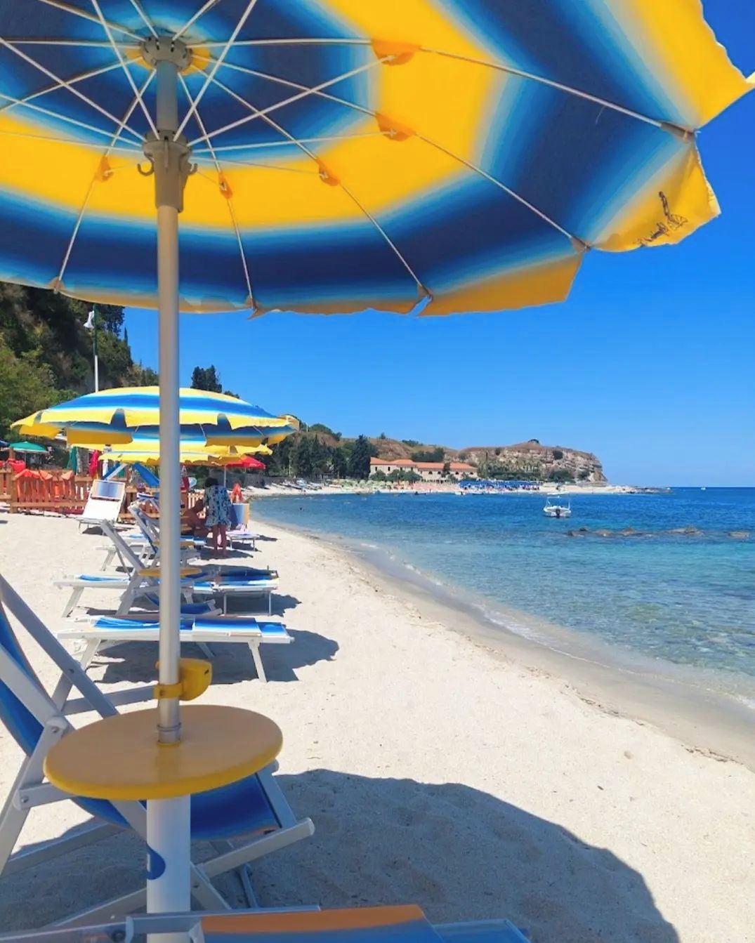Een strand met ligbedden en parasols, helderblauw water en een veraf gelegen rotsachtige kustlijn.