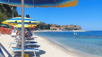 Een strand met ligbedden en parasols, helderblauw water en een veraf gelegen rotsachtige kustlijn.