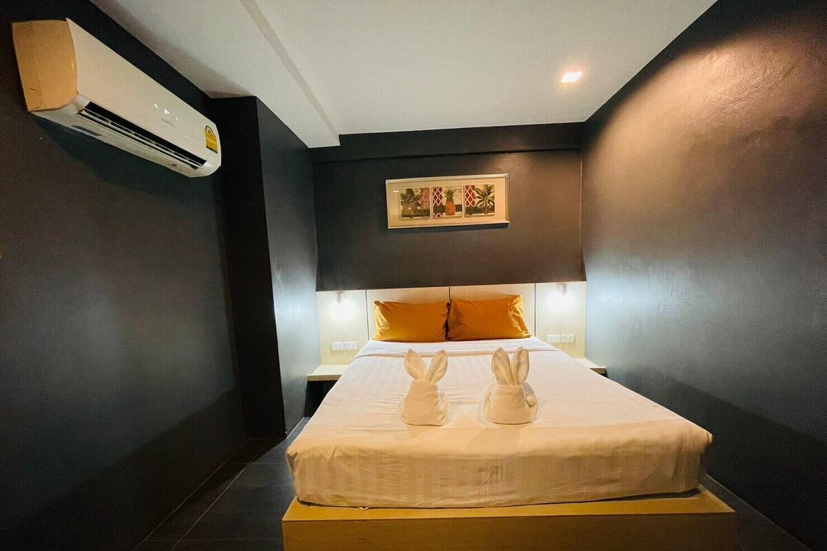 A hotel room with a bed, a nightstand, an air conditioning unit, and a framed picture on the wall.