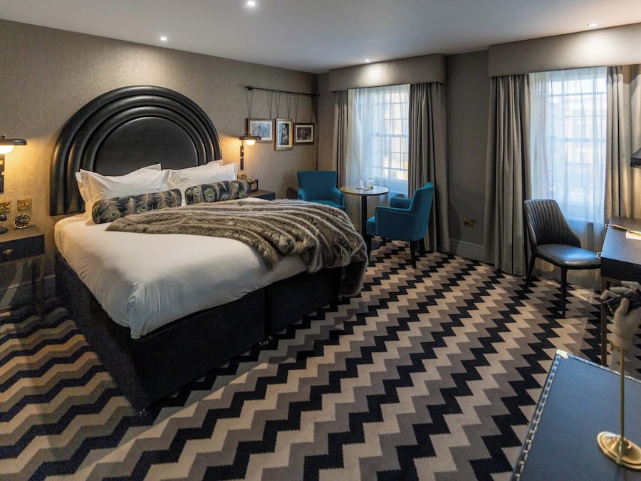 A hotel room with a large bed, a desk, a chair, a small table, and a patterned floor.