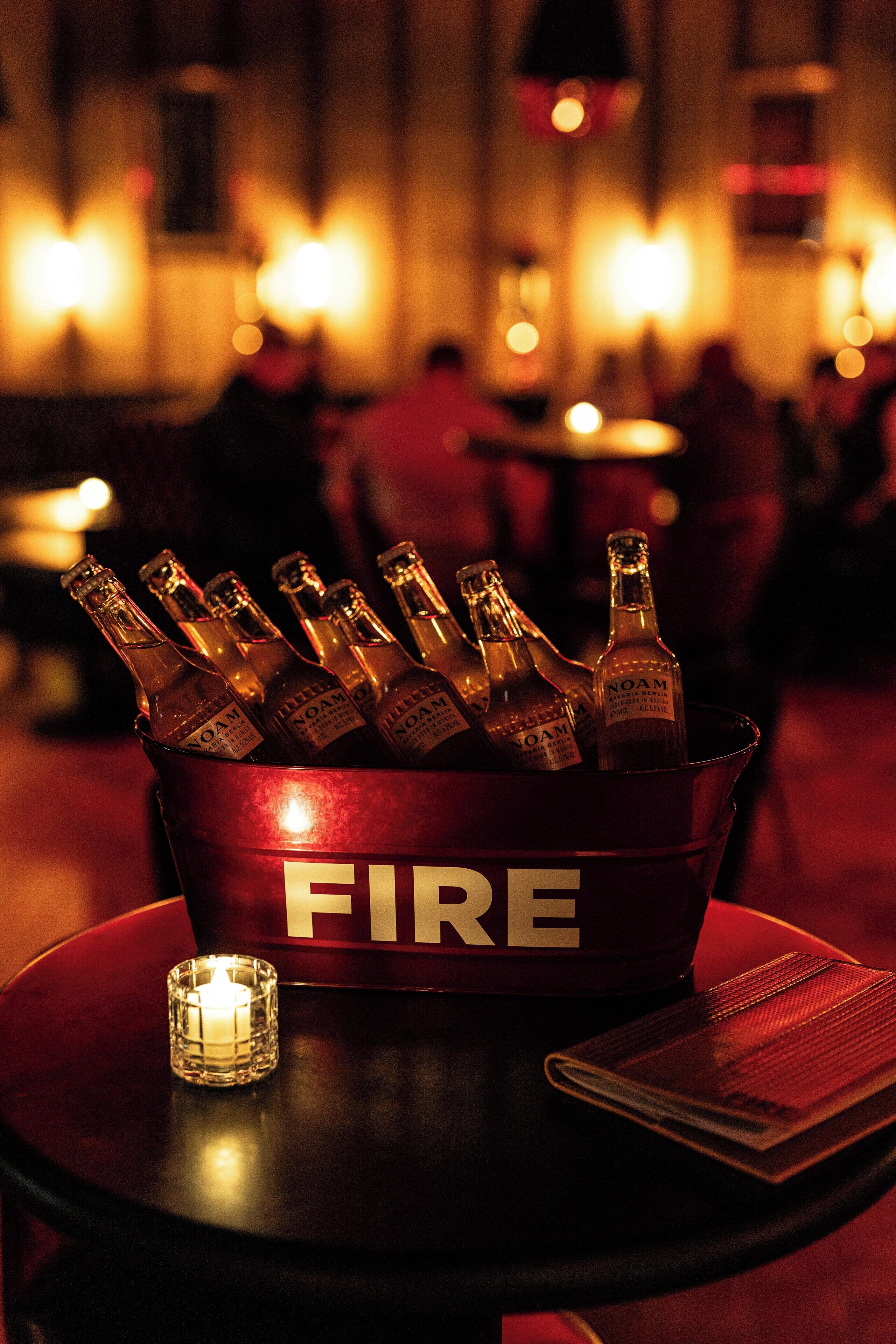 Een emmer met de aanduiding 'FIRE', gevuld met flessen, een brandende kaars en een menukaart op een tafel in een gedimd verlichte omgeving.