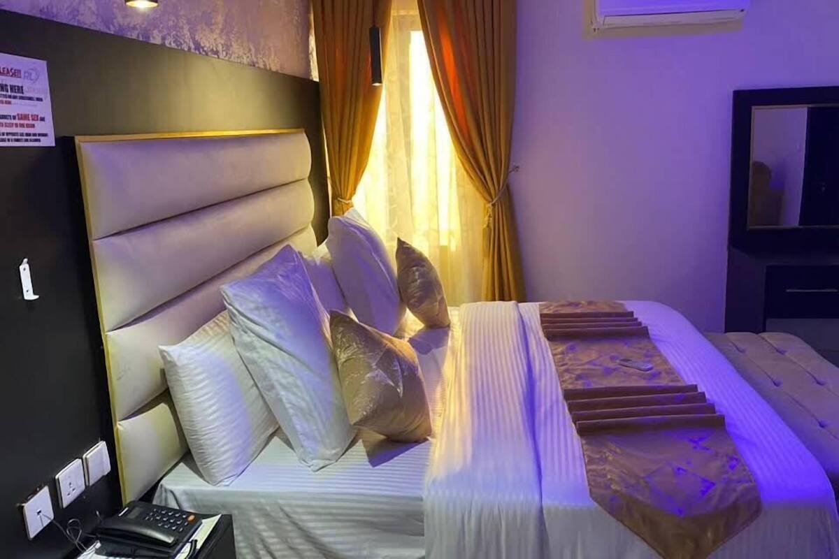 A hotel room with a large bed, a bedside table with a phone and a lamp, a window with curtains, and an air conditioning unit on the wall.