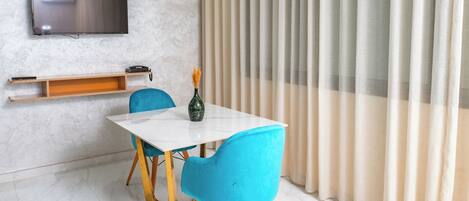 A modern dining area with a white table, two blue chairs, a small shelf with a telephone, and a television mounted on the wall.