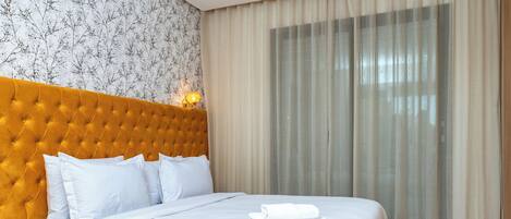 Une chambre d’hôtel avec une tête de lit capitonnée, un lit bien fait avec du linge de lit blanc et une grande fenêtre ornée de rideaux légers.