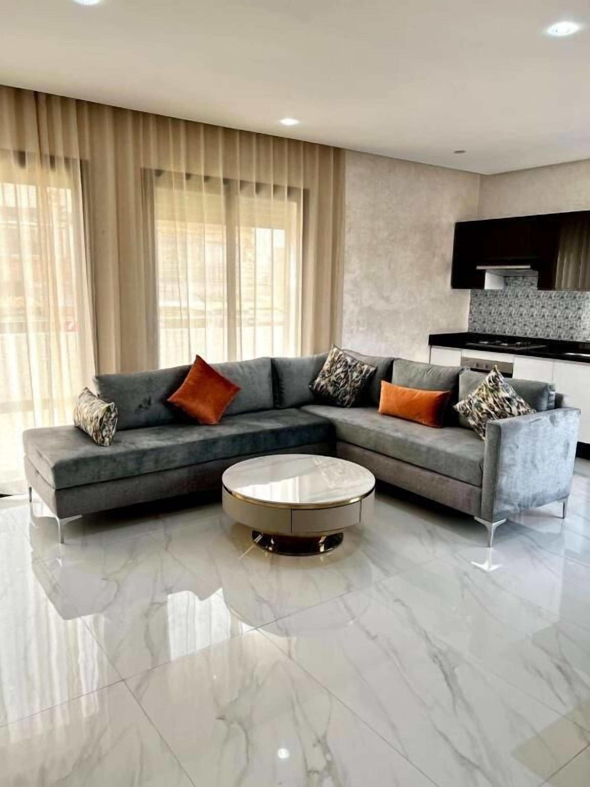 Ein modernes Wohnzimmer mit einem grauen Ecksofa, einem runden Couchtisch und einer Küchenzeile mit schwarzer Arbeitsplatte und Edelstahlgeräten.