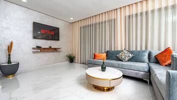 A modern living room with a flat-screen TV displaying 'NETFLIX', a grey sofa with orange and multicolored pillows, a round coffee table with a vase, and a large window with vertical blinds.