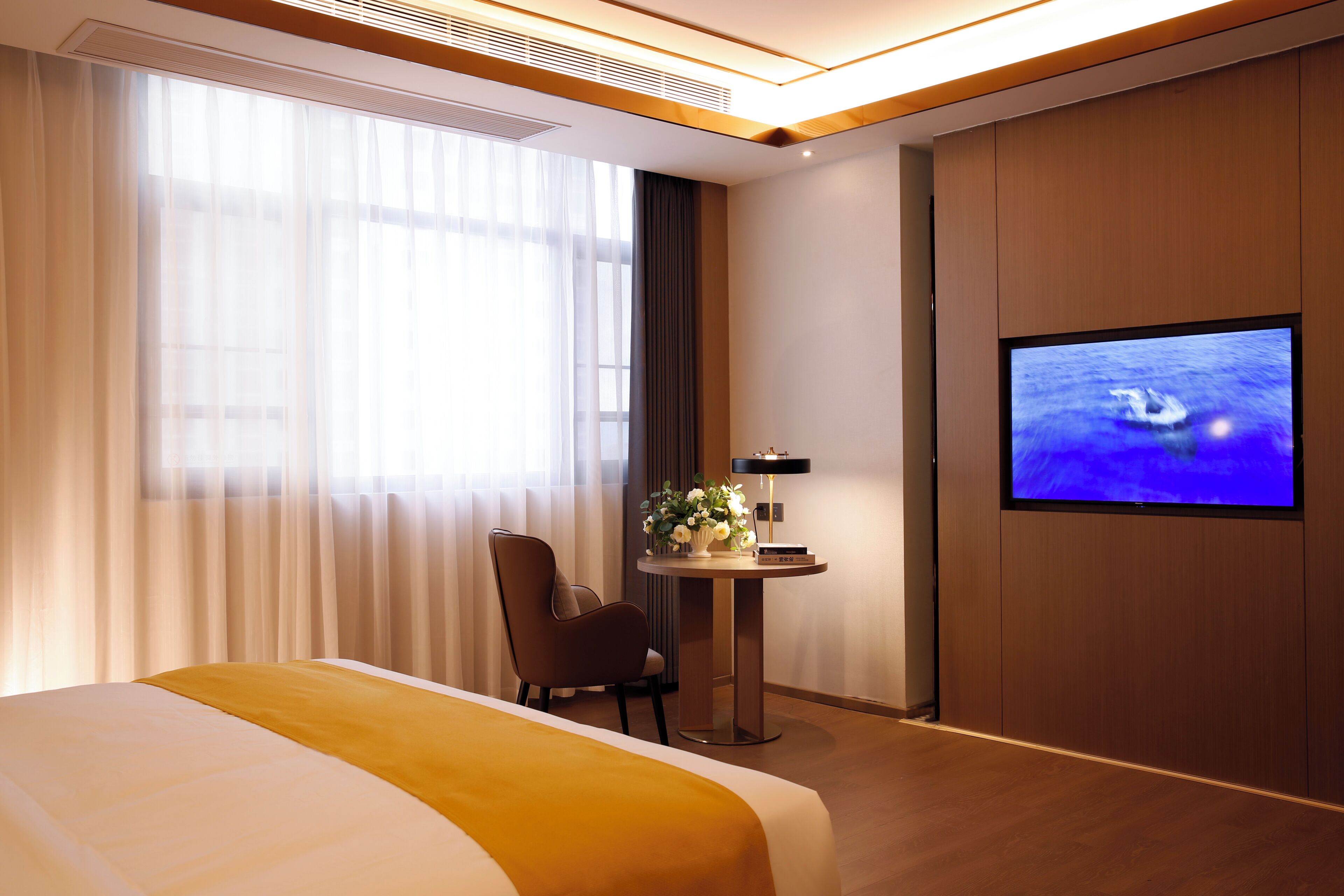 A hotel room with a bed, a TV, a small table with a vase of flowers, and a chair.