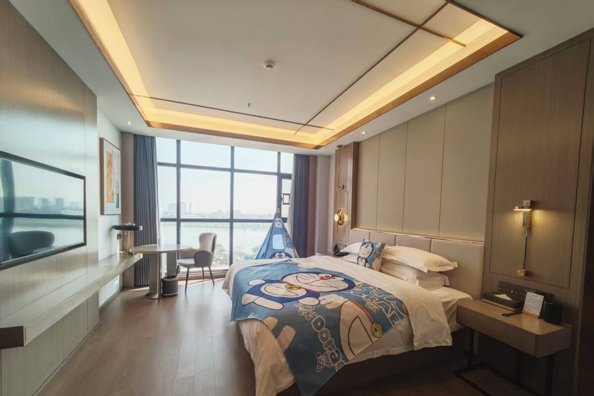 A modern hotel room with a large bed, a desk, and a view of a cityscape through large windows.