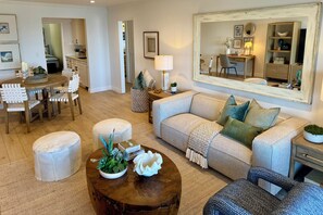 A spacious living room with a sofa, ottomans, a coffee table with plants, a dining area with chairs, and a kitchen in the background.