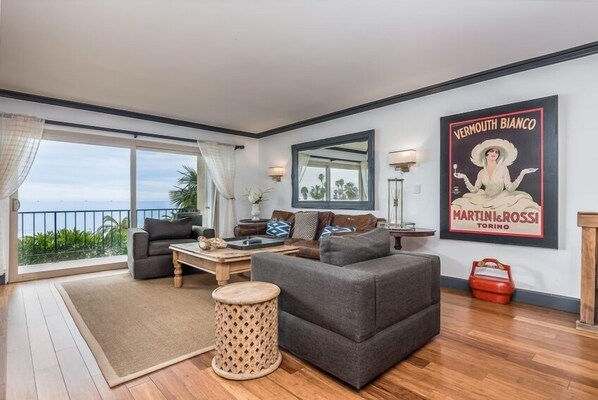 Een woonkamer met een bank, salontafel en een rond houten krukje. Er is een groot raam met uitzicht op palmbomen en een balkon. Een poster aan de muur vermeldt: “Vermouth Bianco Martini & Rossi Torino”.