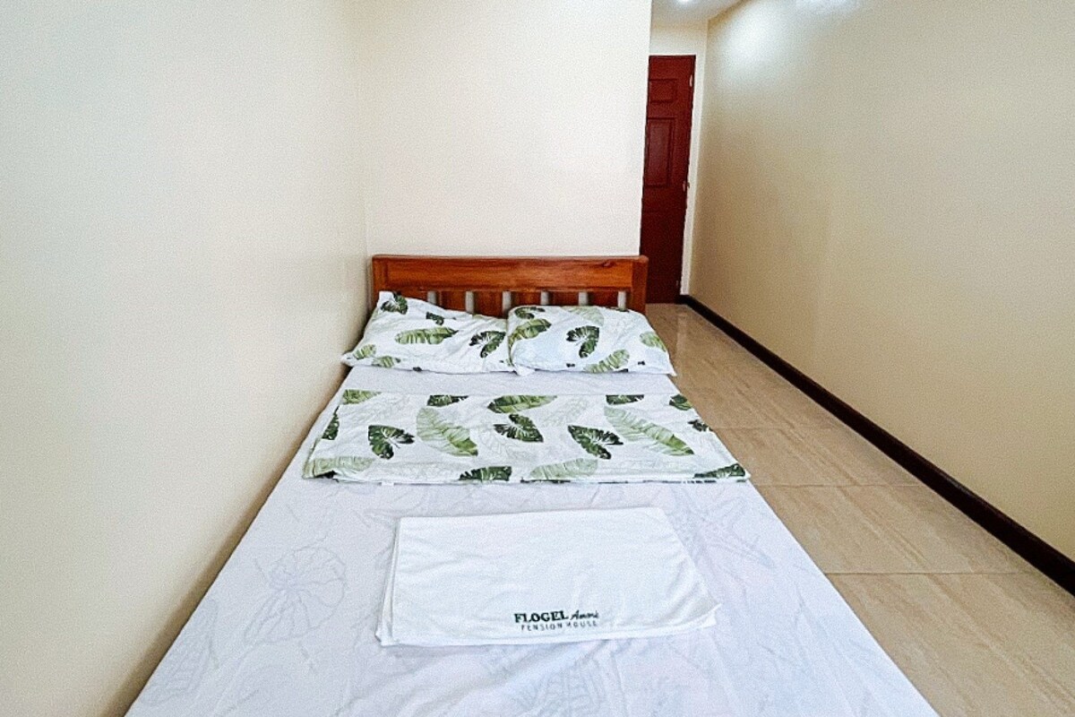 Una cama pequeña y bien hecha, con colchón blanco y ropa de cama estampada con hojas verdes.