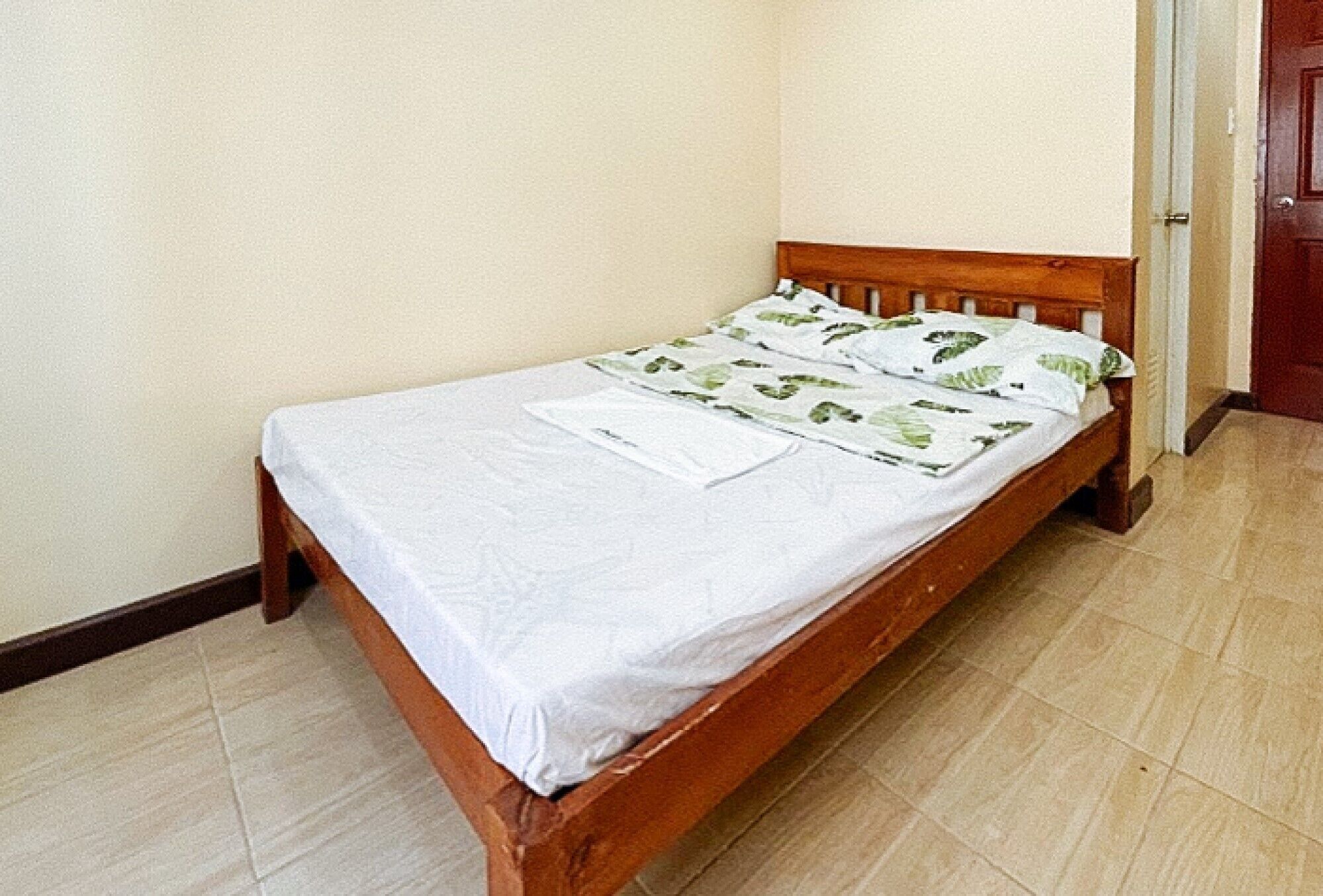 Una cama individual con estructura de madera y ropa de cama blanca, ubicada frente a una pared lisa con una puerta a la derecha.