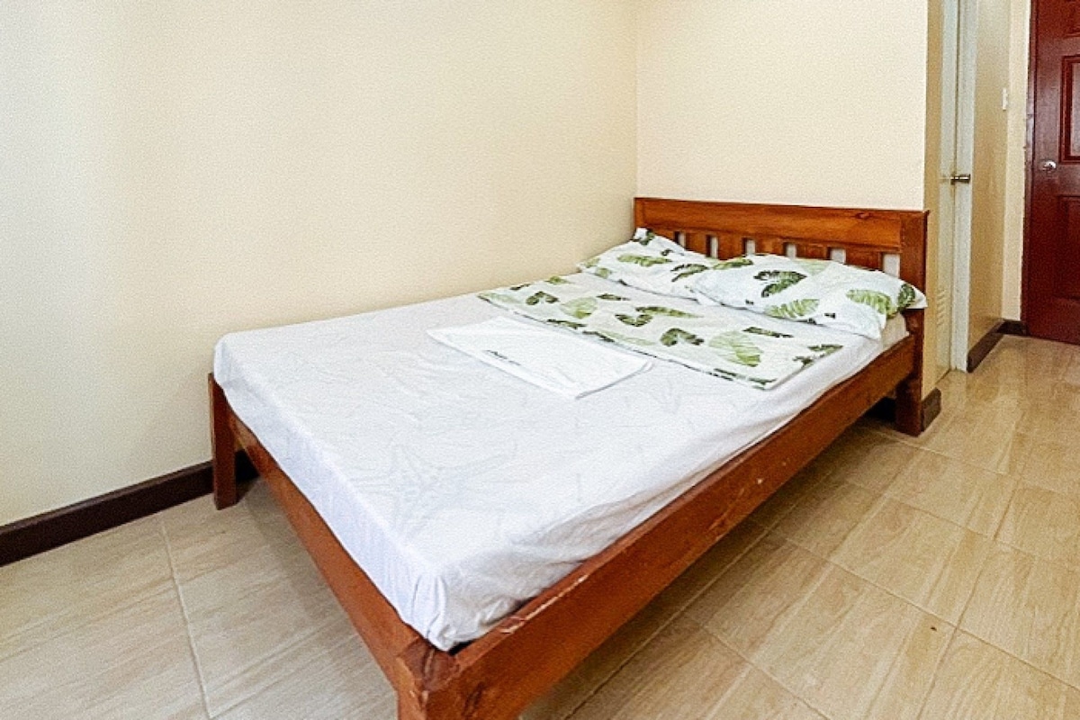 A single bed with a wooden frame and white bedding, positioned against a plain wall with a door to the right.