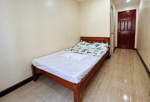Una habitación pequeña con una cama individual, cabecera de madera, colcha blanca y una puerta al fondo.