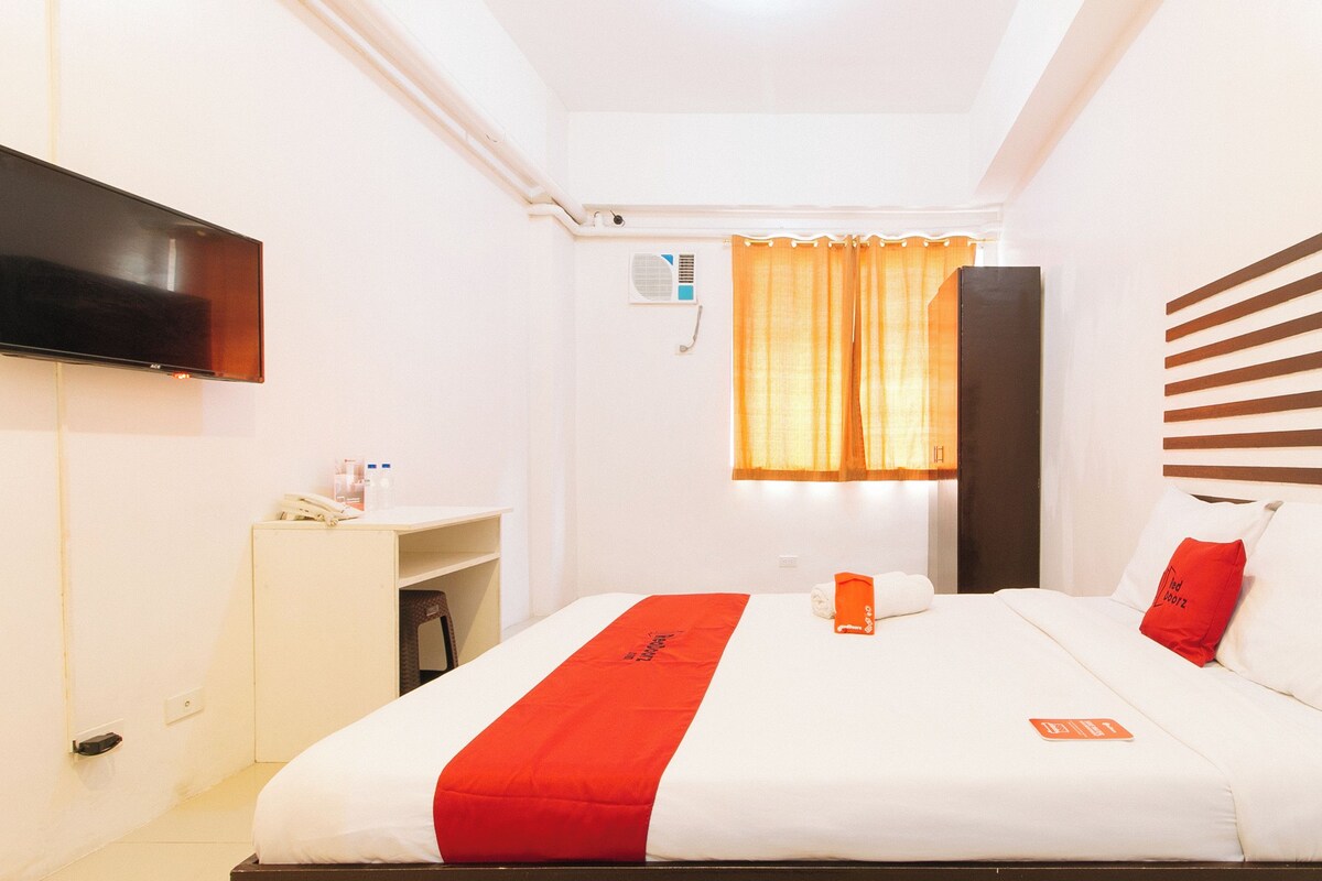 Una habitación de hotel con cama, televisor montado en la pared, escritorio con teléfono y ventana con cortinas.