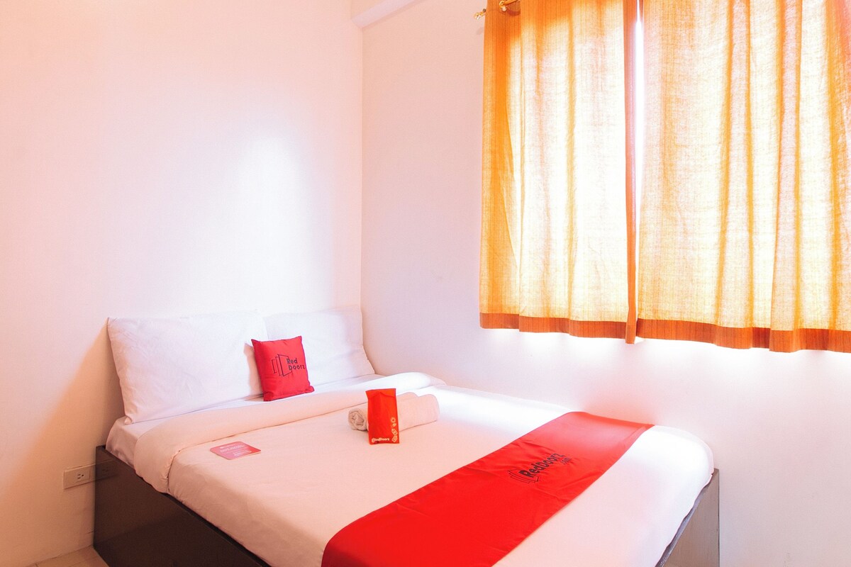 Una habitación de hotel con una cama, un almohadón rojo, una colcha roja y una ventana con cortinas naranjas.