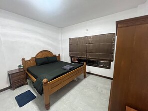 A bedroom with a wooden bed, a small dresser, a window with blinds, and a door.