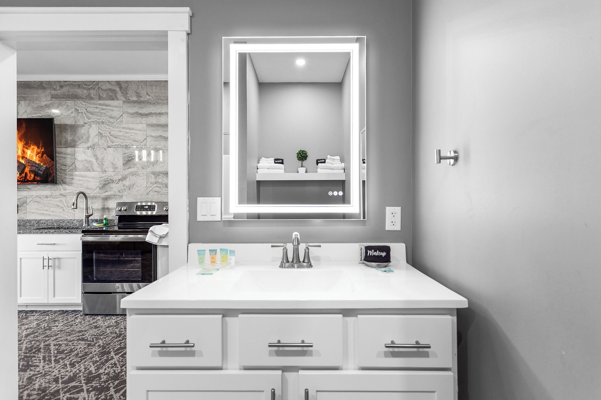 A modern bathroom with a large mirror, white vanity, and a fireplace in the background.
