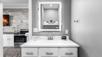 A modern bathroom with a large mirror, white vanity, and a fireplace in the background.