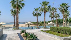 Un espace piscine d’un complexe hôtelier, avec des palmiers, une vue sur la plage et un ciel dégagé.