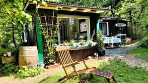 Un espace extérieur rustique aménagé avec un banc en bois, une petite structure en bois portant l’inscription « NATURCAMP », et un jardin avec des plantes en pot et un tonneau en bois.