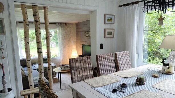 Un coin repas avec une table dressée pour quatre personnes, entourée de chaises en osier, et une vue sur un jardin par la fenêtre.