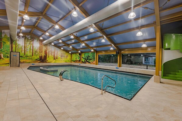 An indoor swimming pool with a tiled floor, metal pool ladders, and a ceiling with wooden beams and hanging lights.