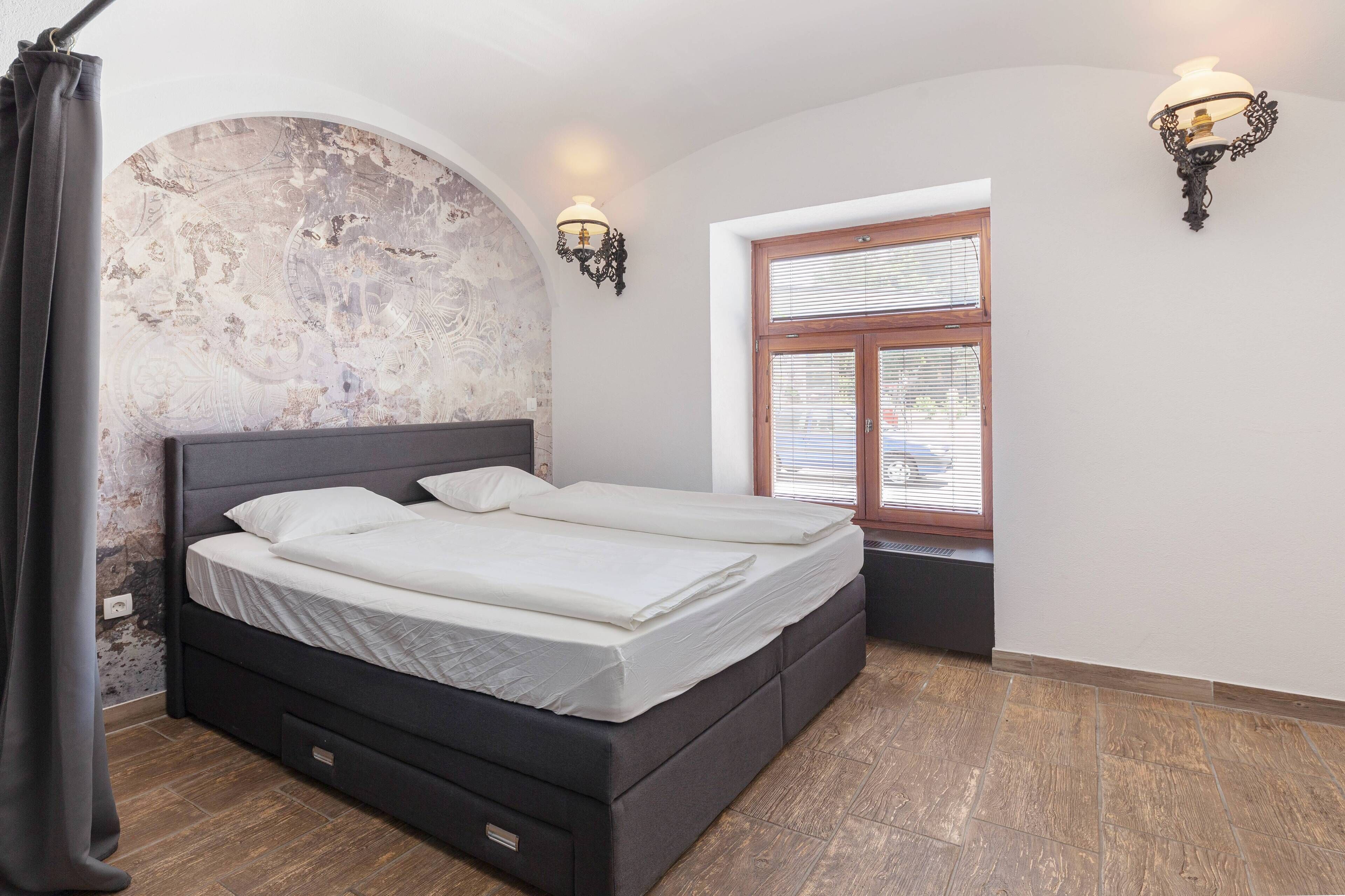 A bedroom with a large bed, two wall sconces, a window with blinds, and a textured wall.