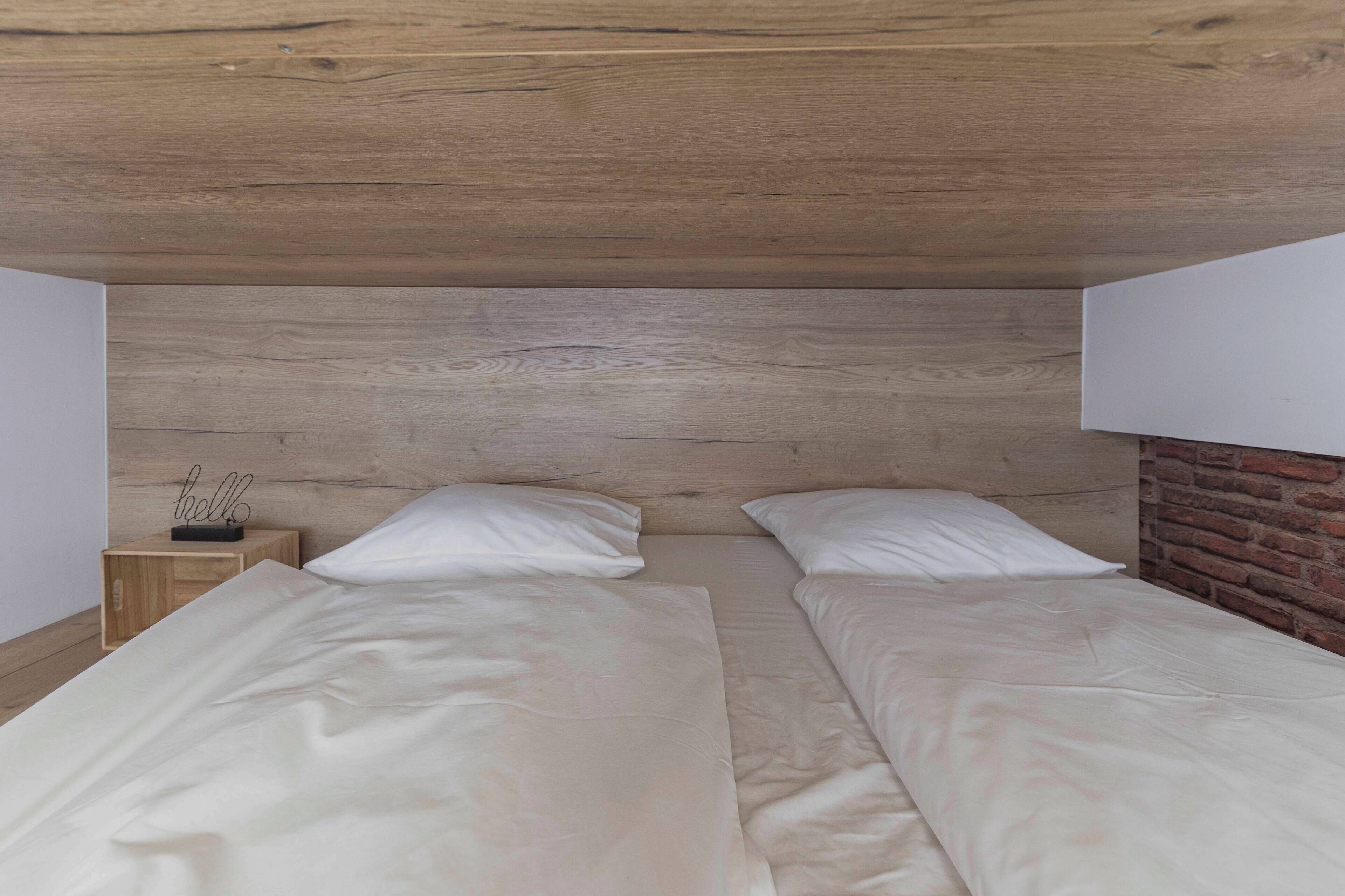 A double bed with white linens in a room with a wooden ceiling and a brick wall.