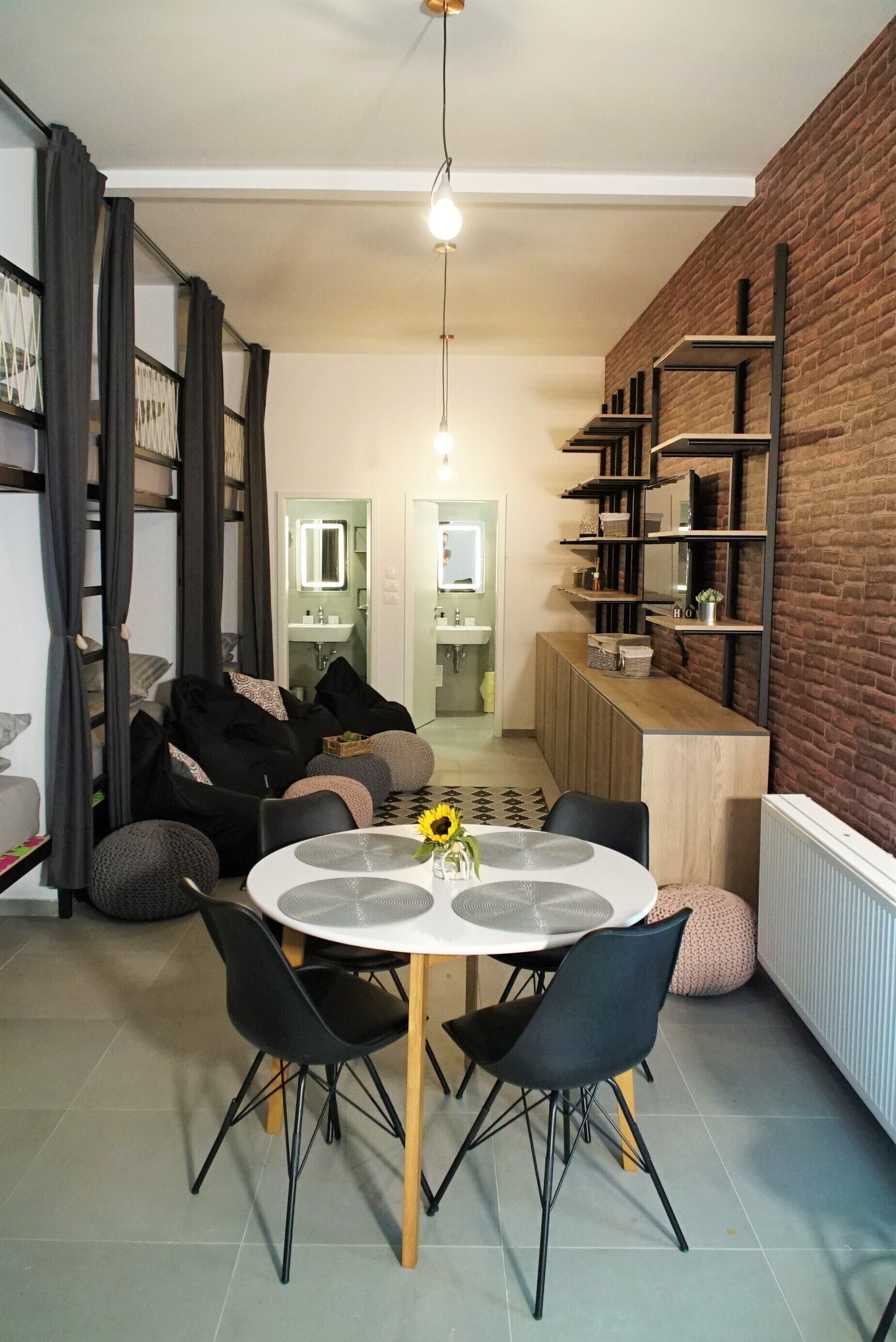 Un espace repas moderne avec une table ronde, des chaises noires et un vase rempli de tournesols. Un mur de briques se trouve sur un côté, et on aperçoit un coin cuisine au fond.
