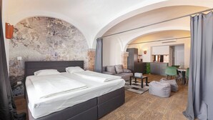 Camera d'albergo moderna con un letto grande, un divano, una scrivania e un angolo cottura visibile attraverso un arco.