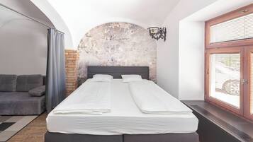 Un dormitorio con cama, sofá gris, pared de ladrillo y ventana con persianas.