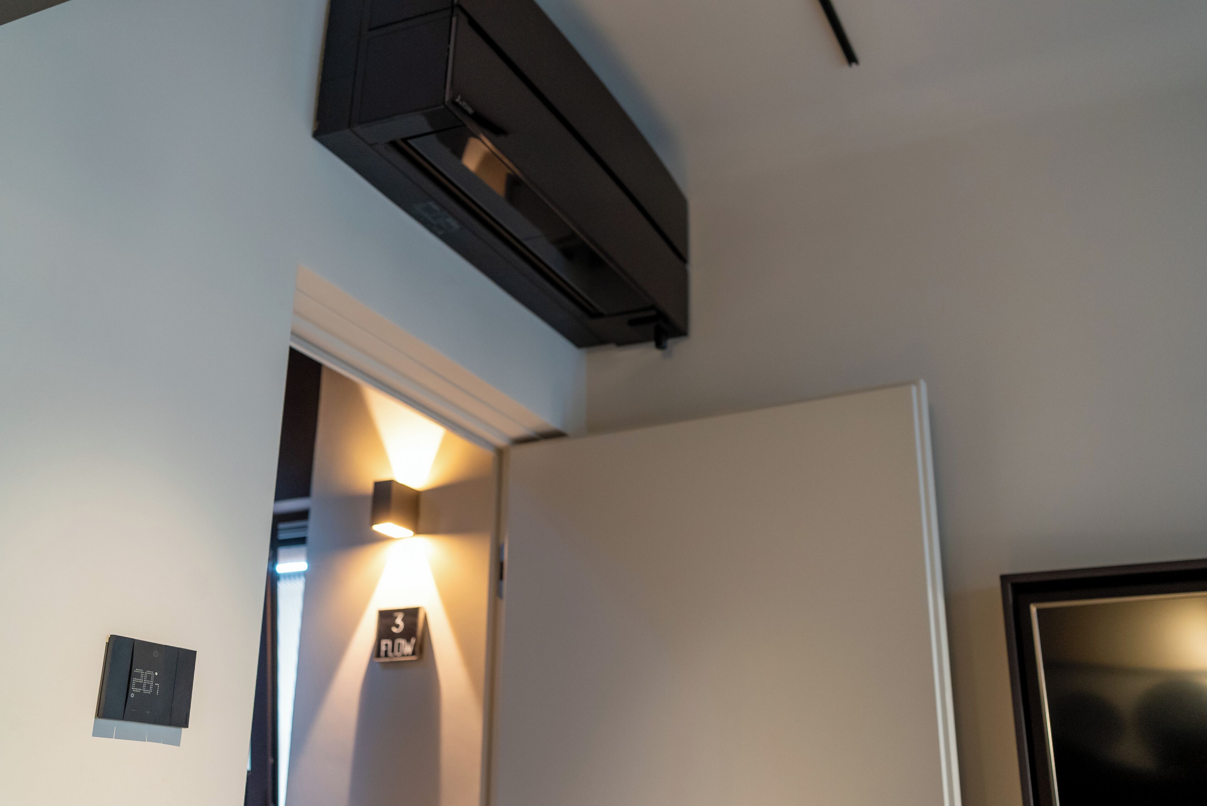 A room with a ceiling-mounted air conditioning unit, a wall-mounted light fixture, and a door.