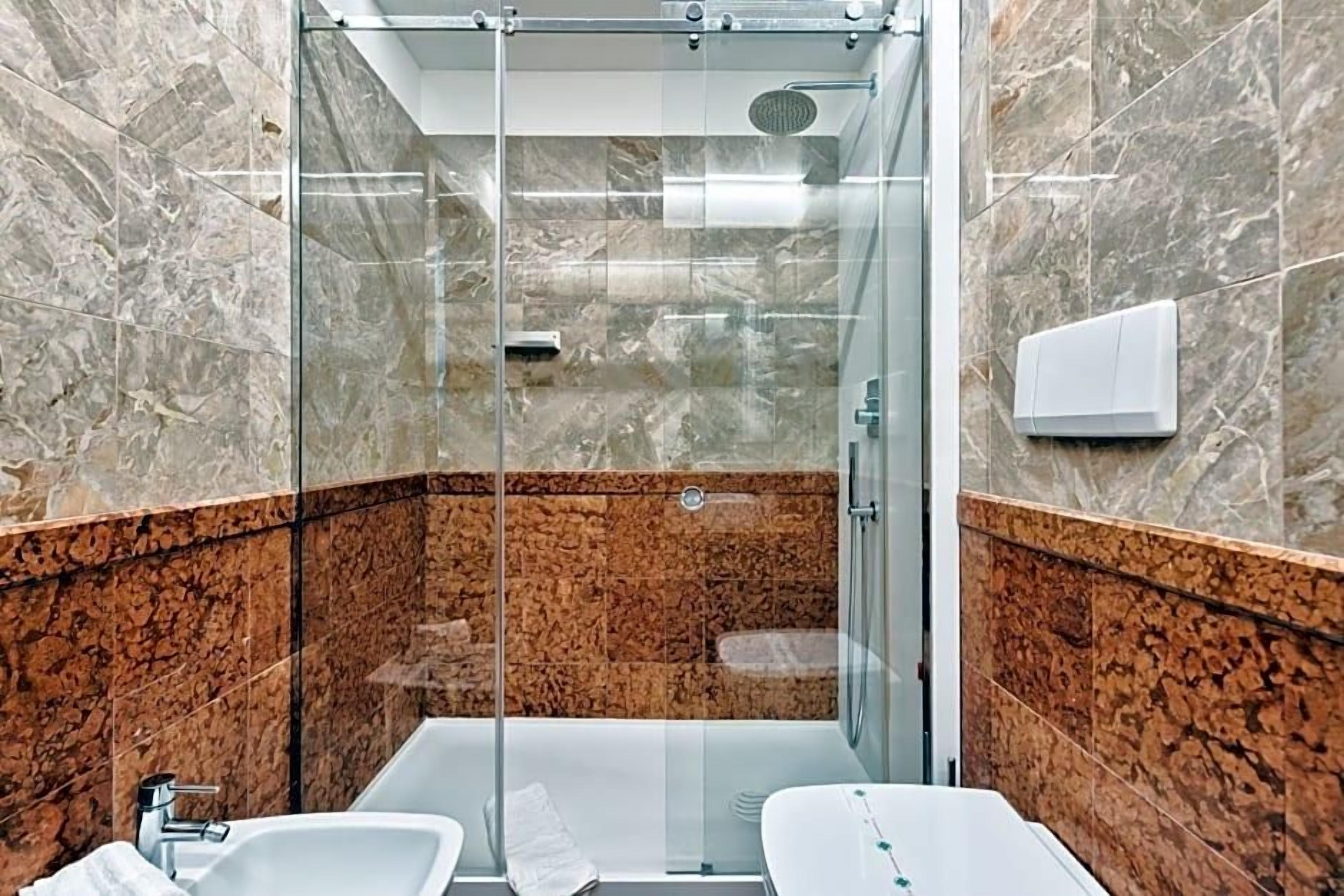 Ein modernes Badezimmer mit einer gläsernen Duschkabine, zwei Waschbecken und einer Wand aus Marmorfliesen.