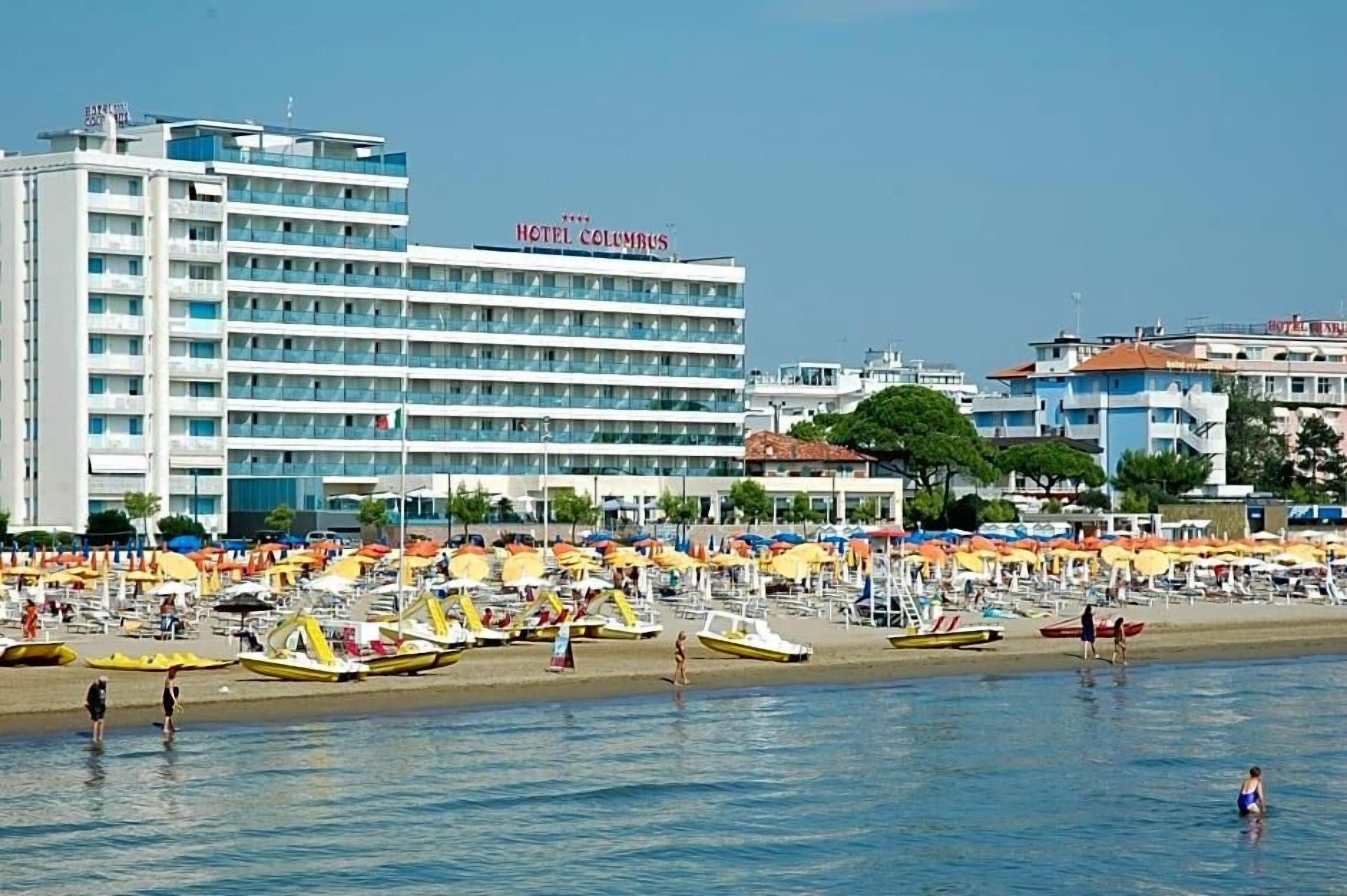 Un hotel con varios pisos y balcones, una playa con numerosas sombrillas y tumbonas, y una vista despejada del mar.