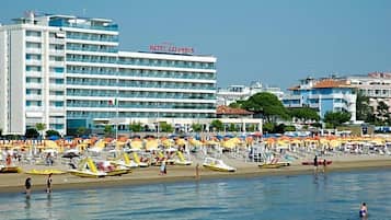 Een hotel met meerdere verdiepingen en balkons, een strand met tal van parasols en ligbedden, en een helder uitzicht op zee.