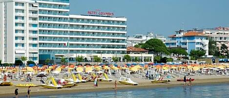 A hotel with multiple floors and balconies, a beach with numerous umbrellas and sun loungers, and a clear view of the sea.