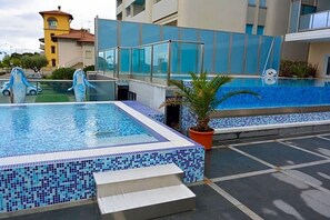A rooftop pool with clear blue water, surrounded by modern buildings and a palm tree. - Hotel Columbus (Lignano Sabbiadoro)