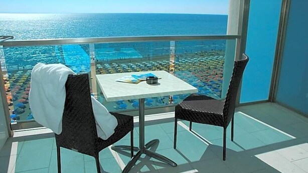 A balcony with a table and chairs, overlooking a pool and beach area. - Hotel Columbus (Lignano Sabbiadoro)