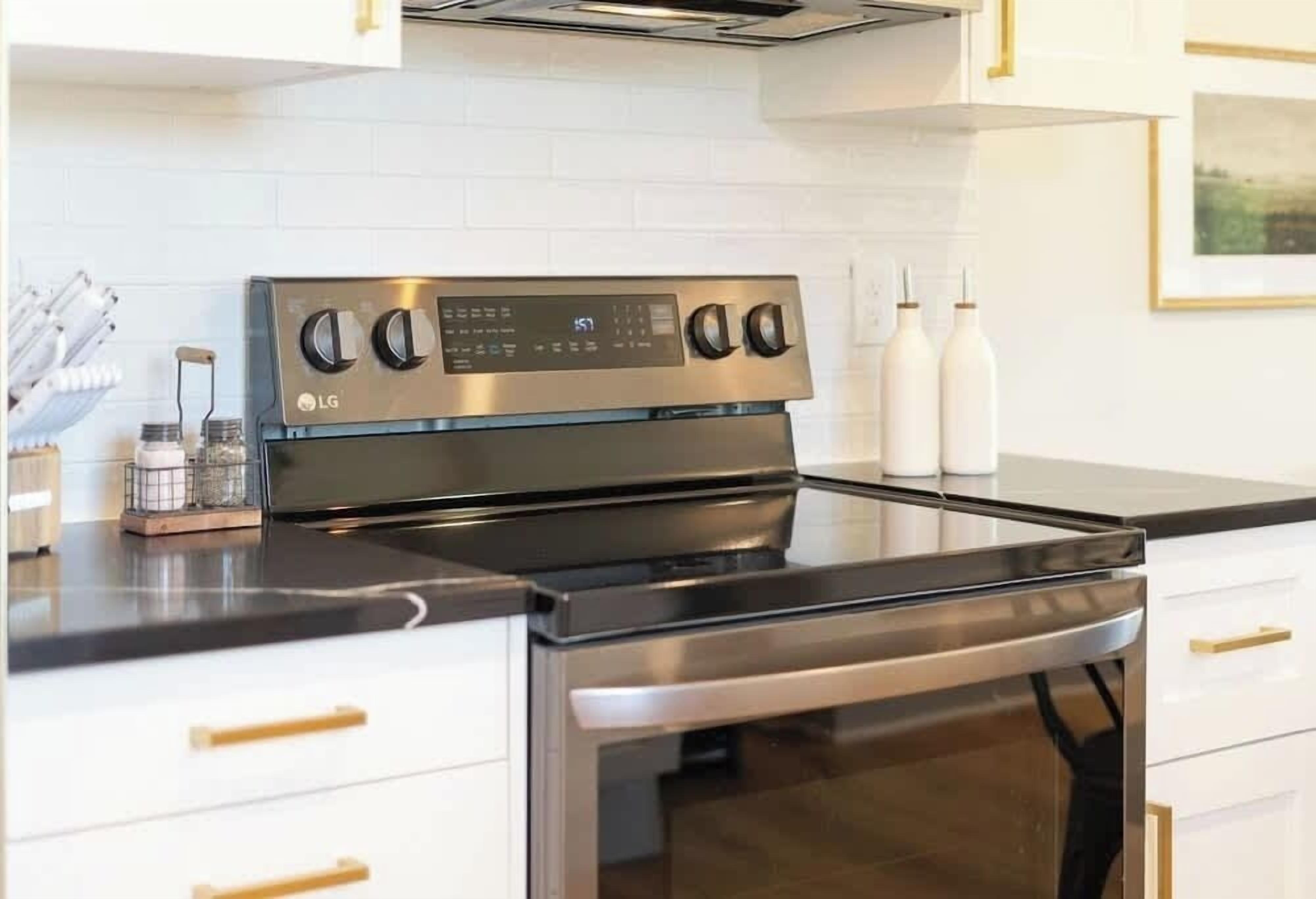 Eine moderne Küche mit einem Edelstahlherd, Backofen und einer schwarzen Arbeitsplatte. Es gibt weiße Schränke mit goldenen Griffen und einer weißen, gefliesten Rückwand.