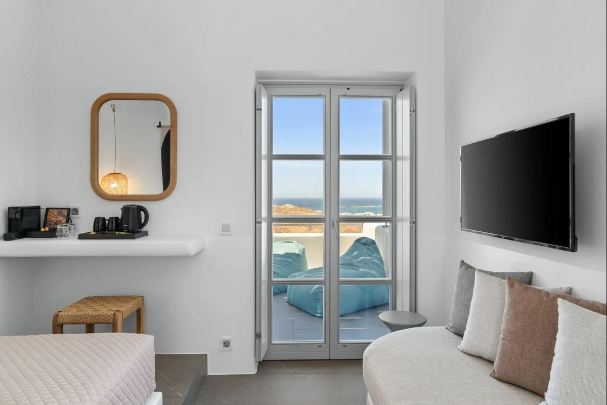 Un dormitorio moderno con cama, una pequeña zona de estar, televisión, espejo y vista al mar a través de un ventanal.