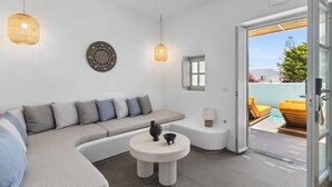 A modern living room with a sectional sofa, a round coffee table, and a glass door leading to an outdoor seating area. - Mr & Mrs White Paros Suites & More (Paros)
