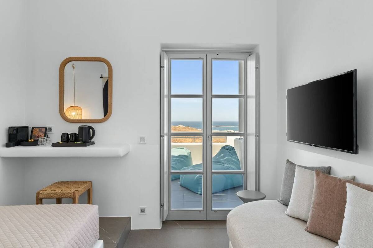 A modern bedroom with a bed, a small seating area, a television, a mirror, and a view of the sea through a glass door.